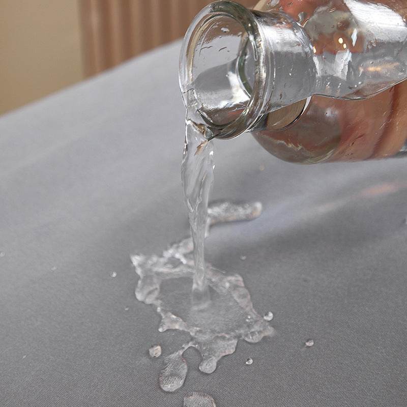 Clear liquid being poured from a glass container onto the surface of the Cozee Comfort Guard Slipcover, demonstrating its water-resistant protection against spills and everyday messes