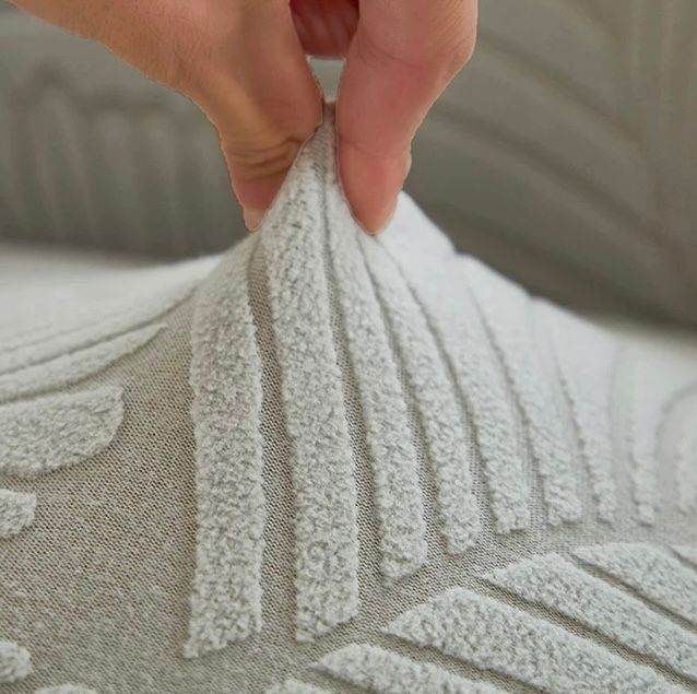 Close-up of hand pinching ultra-soft velvet fabric on the SoftWeave chair cover, showing its flexible stretch and plush texture with embossed leaf detail