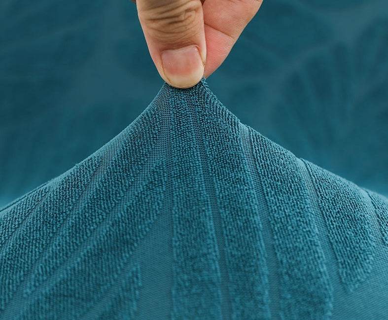 Close-up of a hand holding teal blue textured fabric against a matching blue background, showcasing the detailed weave and flexible stretch of the Cozee SoftWeave Skirted Chair Cover