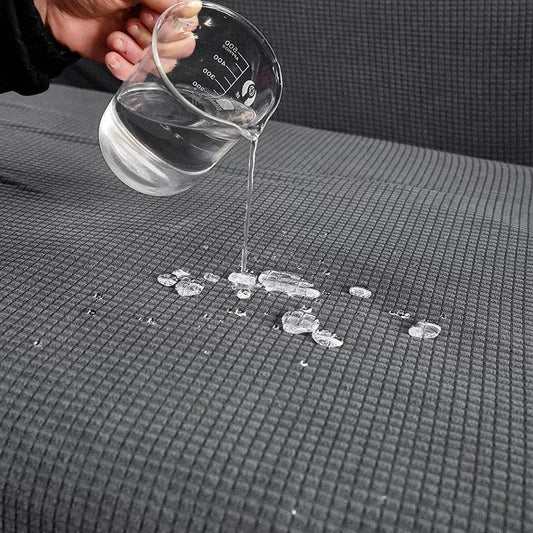 Water being poured from a clear container onto the textured surface of the Cozee Timeless Slipcover, demonstrating its water-resistant fabric that guards against spills and daily messes